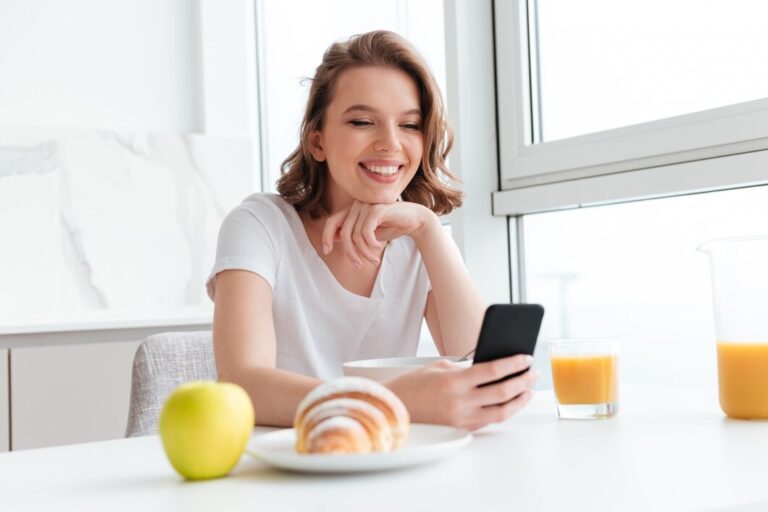 close-up-portrait-happy-brunette-woman-using-mobile-phone-while-having-breakfast-white-kitchen_171337-13149