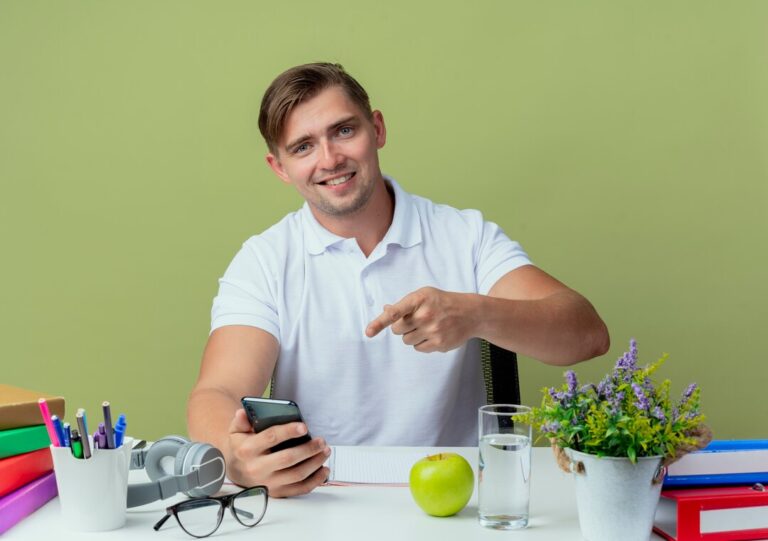 smiling-young-handsome-male-student-sitting-desk-with-school-tools-holding-points-phone-isolated-olive-green_141793-83897